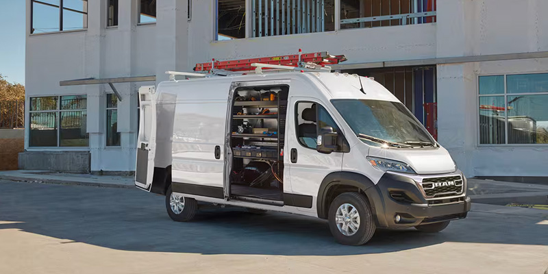 A white Ram cargo van with the side door open, showing its storage compartments, parked in front of a construction site.