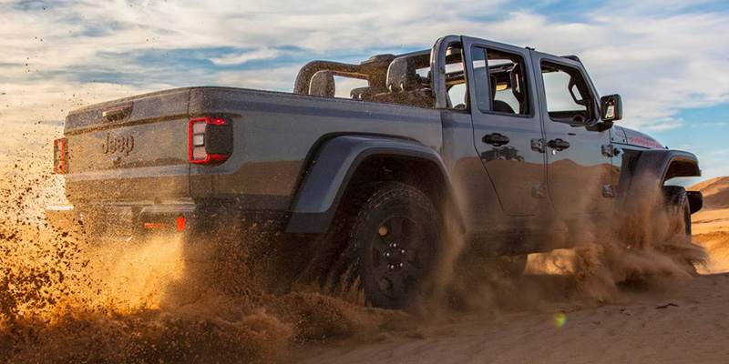 A Jeep Gladiator kicking up sand while driving through desert dunes.